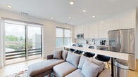 This contemporary single-floor residence presents a clean, modern aesthetic throughout, with crisp white walls, light hardwood flooring, and recessed lighting that create a bright and cohesive atmosphere. The open-plan living and dining area flows seamlessly into a well-equipped kitchen, which features white shaker cabinetry extending to the ceiling, white subway tile backsplash, stainless steel appliances including a range and over-the-range microwave, a matte black faucet, and a white countertop island with bar seating. Large windows and a sliding glass door in the living area fill the space with natural light and provide access to an outdoor balcony with urban views. A wall-mounted flat-screen TV and a colorful area rug anchor the living room, which is finished with light wood floors. The bedrooms are serene and minimal, featuring the same hardwood flooring, white walls, natural light from windows, and built-in overhead storage in select rooms with stylish sconce lighting. The bathrooms maintain the home's modern palette with fully white-tiled walk-in showers enclosed by sleek black-framed glass panels, matte black hardware accents, and clean, minimalist fixtures throughout. The overall design is cohesive, functional, and thoughtfully finished.