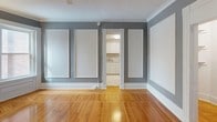 This single-floor space showcases a thoughtful blend of classic architectural detailing and functional design. Throughout the living and sleeping areas, warm-toned hardwood floors with a parquet-style pattern create a welcoming foundation, complemented by gray-painted walls adorned with raised white rectangular panel moldings and crisp white crown molding along the ceiling perimeter. These decorative elements lend a refined, traditional character to the interior.

The main room features a curved bay window area that introduces natural light, with a classic cast-iron radiator positioned beneath it. Additional wall sconces and a ceiling fan contribute to both comfort and ambiance. A closet and secondary doorway are accessible from this room, adding practical storage.

The kitchen is finished in clean white cabinetry with a straightforward, efficient layout visible through a columned doorway from the main living space. Adjacent to the kitchen, the bathroom continues the classic aesthetic with black-and-white octagonal tile flooring, white subway tile walls, a cast-iron radiator, and a shower-tub combination fitted with a patterned curtain. Together, these spaces present a cohesive, classically styled interior with enduring architectural charm.