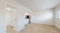 This single-floor unit offers a clean, efficiently designed layout finished consistently in neutral tones and warm-hued tile flooring throughout. Entry is made through a narrow hallway that leads directly into the main living space, a bright and open room featuring two windows fitted with horizontal blinds that allow ample natural light. A wall-mounted mini-split air conditioning unit serves the space for climate control.

The kitchenette is neatly tucked into an alcove off the main room and is well-appointed with white cabinetry featuring black hardware, a speckled countertop, a stainless steel single-basin sink with a matte black faucet, an electric coil range with an overhead exhaust hood, and a stainless steel refrigerator. A secondary hallway connects the living area to the bathroom and provides additional interior access.

The bathroom includes a full tub-and-shower combination with a white surround, matte black fixtures including the faucet, showerhead, and towel bars, and the same tile flooring found throughout the unit. The overall aesthetic is clean and consistent, with white walls, white trim, and black hardware accents unifying the space.