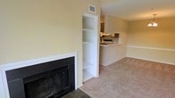 This single-floor space offers a cohesive open-concept layout that connects the living area to the kitchen, creating a functional and comfortable flow throughout. The living area is anchored by a classic fireplace with a clean white surround and dark tile hearth, serving as a natural focal point within the room. Adjacent to the fireplace, a built-in shelving unit with multiple tiers provides convenient display and storage space, seamlessly integrated into the wall. The flooring throughout the living area consists of neutral-toned carpet, contributing to a warm and understated aesthetic. Warm yellow-toned walls complement the light interior, enhancing the brightness of the space. A pass-through countertop connects the living area to the kitchen, which features cabinetry and overhead ventilation, allowing for easy interaction between the two spaces while maintaining a degree of separation. HVAC vents are visible in the ceiling, indicating a central air system. The overall design is straightforward and practical, with architectural details such as the built-in shelving and fireplace adding character to an otherwise clean and neutral interior.