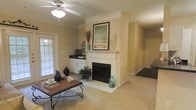 This single-floor space centers around a well-appointed living area with a classic white-mantled fireplace as its focal point. The fireplace features a tiled surround and hearth, topped with a decorative coastal-themed framed artwork, candles, and accent pieces that contribute to a relaxed, inviting ambiance. Neutral beige walls and wall-to-wall carpeting create a warm, cohesive feel throughout the space. A ceiling fan with integrated lighting fixture provides both comfort and illumination. Natural light filters in through a window fitted with white horizontal blinds, brightening the room without overwhelming it. A wooden coffee table with ornate scrollwork legs anchors the seating area, complemented by decorative vases and coastal accents. Adjacent to the living area, a built-in cabinetry section with white lower cabinets and a dark countertop offers additional storage and display space. A hallway visible toward the rear of the space leads to additional rooms, suggesting a practical and straightforward floor plan. Overall, the space presents a comfortable, casually elegant environment with consistent neutral tones and thoughtful decorative touches.
