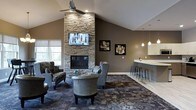 This well-appointed single-floor space is designed with both comfort and style in mind. At its heart stands a dramatic floor-to-ceiling stacked-stone fireplace, serving as the focal point of the main lounge area. A flat-screen television is mounted above the fireplace, making this an ideal setting for both casual conversation and entertainment. The vaulted ceiling adds a sense of openness and grandeur, while a ceiling fan ensures year-round comfort. Warm, taupe-toned walls complement the natural stone textures throughout, creating a cohesive and inviting ambiance. The seating area is furnished with plush armchairs and accent chairs arranged atop a richly patterned area rug, encouraging relaxed gatherings. Large windows allow natural light to filter in, enhancing the welcoming atmosphere. Transitioning to one side of the space, a sleek bar counter with modern pendant lighting and industrial-style bar stools offers a casual spot for socializing. The flooring shifts from patterned carpet in the lounge zone to smooth light-toned flooring near the bar, clearly delineating each area. Decorative framed artwork adorns the walls, adding a finishing touch to this thoughtfully designed communal space.