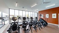 This contemporary fitness center is designed to provide an motivating and fully equipped workout environment. The space is anchored by a striking wall of floor-to-ceiling windows that floods the interior with natural light while offering open views of the surrounding exterior, creating an airy and expansive atmosphere that enhances the exercise experience.

The cardio floor is generously outfitted with a comprehensive selection of equipment, including elliptical machines, treadmills, StairMaster steppers, and stationary bikes, all arranged to maximize usability and flow throughout the space. The warm-toned wood-style flooring contrasts with a bold terracotta accent wall, adding visual character and energy to the room.

Overhead, linear fluorescent lighting runs the length of the ceiling, ensuring the space remains bright and well-illuminated at all times, complementing the natural light from the expansive windows. The clean white ceiling and modern finishes give the facility a polished, professional appearance. The layout is thoughtfully organized to allow for comfortable movement between equipment, making this fitness center a well-rounded and inviting space for daily exercise.