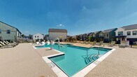 Located at 7801 FM-2920 E in Spring, Texas, this outdoor community pool facility offers a well-appointed, resort-style aquatic amenity space designed for relaxation and recreation. The centerpiece is a generously sized swimming pool with crystal-clear blue water, complemented by a shallow wading area featuring in-water lounge chairs — ideal for sunbathing while staying cool. An adjacent hot tub/spa provides a dedicated relaxation zone, while wide entry steps with handrails offer easy pool access. The pool is also equipped with an ADA-compliant lift, ensuring accessibility for all users. Surrounding the pool is an expansive concrete sun deck finished with a textured, slip-resistant surface, furnished with numerous chaise lounges and shaded seating areas with umbrellas. A single-story clubhouse structure anchors one end of the space, featuring a classic farmhouse-style exterior with board-and-batten siding, a pitched roof, and large green-framed doors. The entire pool area is enclosed by wrought iron fencing, providing security while maintaining open views of the surrounding residential community. The space benefits from abundant natural sunlight under open Texas skies, creating a bright and inviting outdoor atmosphere year-round.