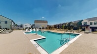 Located at 7801 FM-2920 E in Spring, Texas, this outdoor community pool facility offers a well-appointed, resort-style aquatic amenity space designed for relaxation and recreation. The centerpiece is a generously sized swimming pool with crystal-clear blue water, complemented by a shallow wading area featuring in-water lounge chairs — ideal for sunbathing while staying cool. An adjacent hot tub/spa provides a dedicated relaxation zone, while wide entry steps with handrails offer easy pool access. The pool is also equipped with an ADA-compliant lift, ensuring accessibility for all users. Surrounding the pool is an expansive concrete sun deck finished with a textured, slip-resistant surface, furnished with numerous chaise lounges and shaded seating areas with umbrellas. A single-story clubhouse structure anchors one end of the space, featuring a classic farmhouse-style exterior with board-and-batten siding, a pitched roof, and large green-framed doors. The entire pool area is enclosed by wrought iron fencing, providing security while maintaining open views of the surrounding residential community. The space benefits from abundant natural sunlight under open Texas skies, creating a bright and inviting outdoor atmosphere year-round.