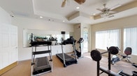 This fitness center is a thoughtfully designed, single-level space offering a comprehensive range of exercise equipment within a bright and open environment. The room features neutral-toned carpeted flooring and crisp white walls, creating a clean and functional atmosphere throughout.

The cardio area is equipped with multiple treadmills, elliptical trainers, and stationary and recumbent bikes, providing a variety of aerobic workout options. A dedicated strength training section includes a multi-station cable machine, a weight bench, a barbell rack, and an assortment of dumbbells and free weights, as well as a stability ball for flexibility and core training. A wall-mounted flat-screen television is positioned for convenient viewing during workouts.

Large wall-length mirrors are strategically placed to visually expand the space and allow exercisers to monitor their form. Two ceiling fans provide consistent air circulation, complementing the recessed lighting overhead. Large windows fitted with vertical blinds allow abundant natural light to filter in while offering views of an outdoor pool area, enhancing the overall ambiance. A glass-panel door provides direct access to the exterior, further connecting the indoor space to its outdoor surroundings.