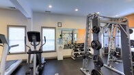 Located at 2230 Greenville Boulevard in Greenville, NC, this fitness center is a well-appointed, single-floor facility designed to support a comprehensive workout experience. The space features a clean, modern aesthetic with white walls, recessed lighting, and dark rubber flooring that provides both durability and comfort underfoot. Natural light filters in through a window, contributing to the bright and energizing atmosphere of the room. A large wall mirror runs along one side of the space, visually expanding the room while allowing users to monitor their form during exercises. The facility is equipped with a range of cardio machines, including an elliptical trainer, positioned to make efficient use of the floor space. The free weight area includes a well-organized dumbbell rack stocked with a full range of weights, alongside a weight plate storage system. A multi-function cable and strength machine anchors the resistance training section, offering versatility for a variety of exercises. A water cooler is conveniently placed within the space, ensuring easy access to hydration. An accent wall in warm orange adds a motivating visual element to the environment. Informational signage is posted on the walls, rounding out this practical and functional fitness facility.