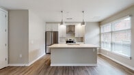 Located at 30 W First Ave in Columbus, Ohio's vibrant Short North area, this single-floor unit offers a clean, contemporary design with an open-concept layout that promotes a seamless flow between living spaces. Warm-toned wood-look plank flooring runs throughout, anchoring the neutral color palette of soft grays and beige that defines the interior.

The kitchen serves as a focal point of the space, featuring white shaker-style cabinetry, a large center island with an integrated sink and crisp white countertops, and stainless steel appliances including a French door refrigerator and range with a wall-mounted hood. Sleek pendant lights hang above the island, adding a modern touch. The kitchen opens directly to the dining and living areas, which are bathed in natural light from multiple windows fitted with horizontal blinds.

The living area is generously proportioned, with architectural details including structural columns and varied ceiling lines that add visual interest. Adjacent bedroom spaces are well-lit by windows with exterior views and maintain the same consistent flooring and neutral tones.

The bathroom is finished with large-format tile flooring, a white vanity with ample storage, a framed mirror, and a walk-in shower. The overall design of the unit is modern and move-in ready, offering a refined yet functional living environment in a prime Columbus location.