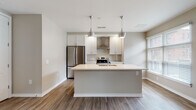 Located at 30 W First Ave in Columbus, Ohio's vibrant Short North area, this single-floor unit offers a clean, contemporary design with an open-concept layout that promotes a seamless flow between living spaces. Warm-toned wood-look plank flooring runs throughout, anchoring the neutral color palette of soft grays and beige that defines the interior.

The kitchen serves as a focal point of the space, featuring white shaker-style cabinetry, a large center island with an integrated sink and crisp white countertops, and stainless steel appliances including a French door refrigerator and range with a wall-mounted hood. Sleek pendant lights hang above the island, adding a modern touch. The kitchen opens directly to the dining and living areas, which are bathed in natural light from multiple windows fitted with horizontal blinds.

The living area is generously proportioned, with architectural details including structural columns and varied ceiling lines that add visual interest. Adjacent bedroom spaces are well-lit by windows with exterior views and maintain the same consistent flooring and neutral tones.

The bathroom is finished with large-format tile flooring, a white vanity with ample storage, a framed mirror, and a walk-in shower. The overall design of the unit is modern and move-in ready, offering a refined yet functional living environment in a prime Columbus location.