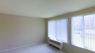 This single-floor space centers around a spacious, open living area finished with neutral beige walls and light-colored carpeting that lends a clean, understated aesthetic throughout. The room is characterized by an abundance of natural light, drawn in through multiple large windows dressed with vertical blinds that allow for flexible light control. The windows offer views of the surrounding outdoor environment, contributing to an airy and open atmosphere.

Practical heating and cooling infrastructure is visibly integrated into the space: baseboard radiator units run beneath the windows along the perimeter, while a wall-mounted air conditioning unit provides additional climate control. The smooth, light-toned ceiling complements the overall neutral palette, maintaining a consistent and cohesive look.

The layout is open and unobstructed, offering versatile potential for furnishing and use. White baseboards frame the carpeted floor cleanly, adding a modest architectural detail. Overall, the space presents a functional and simply designed interior, well-suited for a variety of living arrangements.