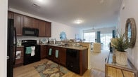 Located at 36916 Crooked Hammock Way in Lewes, Delaware, this single-floor home offers a well-designed, open-concept layout that seamlessly connects the kitchen and living areas. The kitchen is appointed with rich dark wood cabinetry, granite countertops, and black appliances, including a range, microwave, and dishwasher. A peninsula with a double sink serves as a natural divider between the kitchen and the living space, providing both functionality and an open sightline throughout the home. Hardwood-style flooring runs through the kitchen area, complemented by a decorative area rug that adds warmth and character to the space. The living area transitions to plush carpeting and is bathed in natural light from large windows and a sliding glass door, which also provides outdoor access. A ceiling fan keeps the space comfortable year-round. The overall ambiance is bright and airy, with a coastal-inspired décor palette featuring teal and neutral tones that enhance the relaxed, welcoming atmosphere. This thoughtfully designed single-story home offers comfortable, accessible living in a desirable Lewes location.