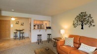 This single-floor space is thoughtfully designed with an open-concept layout that seamlessly connects the main living area, dining space, and kitchen. The living room serves as the central gathering area, featuring carpeted flooring, neutral-toned walls, and warm ambient lighting provided by a floor lamp. A leather sofa anchors the seating area, complemented by a wooden coffee table and decorative wall accents that add character to the space.

A defining architectural feature is the pass-through bar counter that connects the living area to the kitchen, topped with a ledge that accommodates bar stools — ideal for casual dining or conversation. The kitchen, visible through the pass-through window, is equipped with stainless steel appliances, including a refrigerator, and features warm wood-toned cabinetry that adds a cozy contrast to the neutral surroundings.

Adjacent to the living room, the dining area offers additional seating with a bistro-style table and chairs, continuing the open and accessible flow of the space. Throughout, the consistent neutral color palette and soft lighting create a relaxed, welcoming ambiance well-suited for everyday living.