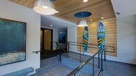 This single-floor space makes a striking first impression with its carefully curated surf-inspired design aesthetic. The entryway sets the tone immediately, featuring a warm wood-paneled ceiling that adds natural texture and depth, complemented by sleek modern pendant lights with metallic dome shades. Recessed lighting throughout ensures the space is well-illuminated at every turn.

A standout feature of the space is the display of vivid, hand-painted surfboards mounted along a slatted wood accent wall. Each board showcases bold, colorful artwork depicting coastal and cityscape scenes, lending the space a dynamic, gallery-like atmosphere. The combination of the wood wall cladding and the artistic surfboards creates a focal point that is both energetic and visually engaging.

The flooring throughout is composed of large-format slate-gray tiles, providing a grounded, contemporary foundation that contrasts beautifully with the warmth of the ceiling. Sleek metal railings guide movement through a multi-level transition area connected by wide steps, adding an architectural element that enhances the sense of flow. A framed nature-inspired painting on the white wall further contributes to the coastal and outdoorsy character of the space, making the overall ambiance feel adventurous, stylish, and welcoming.