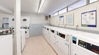 Located at 3020 Glencrest Rd in Burlington, Ontario, this shared laundry facility is a well-organized, single-floor space designed to serve a residential building's laundering needs efficiently. The room is equipped with a substantial row of front-loading commercial washers, complemented by both stacked and standalone dryers positioned along the perimeter walls, providing ample capacity for multiple users simultaneously. The flooring throughout consists of classic small-tile mosaic in neutral tones, offering a durable and easy-to-maintain surface. Fluorescent lighting runs along the ceiling, ensuring bright, consistent illumination across the entire space. High horizontal windows along one wall allow additional natural light to filter in. The walls feature a two-tone finish with a light blue wainscoting detail and white upper walls, lending the room a clean and orderly appearance. A utility sink station is available for pre-treating garments or hand washing, and a paper towel dispenser nearby adds convenience. Informational postings and a bulletin board are mounted on the walls, providing residents with laundry instructions and building notices. A folding table offers a practical surface for sorting and folding laundry. Baseboard heating ensures comfort during cooler months, and a fire alarm system is visible, reflecting attention to safety standards throughout the facility.