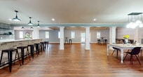 This single-story clubhouse is a well-appointed community gathering space that blends modern farmhouse aesthetics with functional design. Rich hardwood flooring runs throughout, complementing the cool blue-gray walls and crisp white trim that define the interior palette.

The main open hall is expansive, anchored by white decorative columns and outfitted with bar-height tables and black metal stools, making it ideal for events and social gatherings. Natural light filters in through multiple windows, and recessed lighting ensures the space remains bright at all times. A vaulted ceiling with an octagonal tray detail and a statement light fixture adds architectural interest to one section of the hall.

A dedicated lounge area features two tufted leather sofas, a stone-clad fireplace with a wood mantel, a wall-mounted TV, and textured accent wallpaper — creating an inviting, relaxed atmosphere. Adjacent to the lounge is a kitchenette and bar area with a brick-faced counter, patterned tile backsplash, open shelving, and stainless steel appliances. The welcoming entry foyer features a chandelier pendant light, a round pedestal table, and views through glass doors to the outdoors.