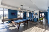 Located at 303 W Blount Ave in Knoxville, Tennessee, this single-floor amenity space is designed with a bold, contemporary aesthetic defined by rich teal and blue walls, warm hardwood-style flooring, and eclectic furnishings throughout. The entrance welcomes visitors with an open foyer featuring large glass doors, abundant natural light, and a seating area with retro-inspired furniture. A striking decorative column wrapped in bold patterned wallpaper anchors the central corridor, adding a playful design element to the space. The lounge area is a standout, anchored by a marble-surround fireplace, a tufted blue sectional sofa, a velvet tufted ottoman, and a statement chandelier with industrial-style cage lighting. Vibrant artwork and chartreuse accent seating add pops of color throughout. The dining and kitchen area features floor-to-ceiling windows with outdoor views, pendant lantern lighting, communal live-edge wood tables, patterned tile flooring, and counter appliances for resident convenience. The game room offers a full-size billiards table, wall-mounted television, and expansive windows that flood the space with natural light. A dedicated business center provides workstations with desktop computers, task lighting, and nature-inspired wall art, creating a focused and functional environment. Together, these areas form a cohesive, energetic communal hub designed for both relaxation and productivity.