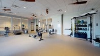 This fitness center at 1100 S Main St in Greenville, South Carolina, offers a comprehensive workout environment thoughtfully designed for a wide range of exercise routines. The space is divided into distinct functional zones, each equipped to support different training needs. The cardio area features multiple treadmills and elliptical machines positioned along large floor-to-ceiling windows that flood the room with natural light and provide open views of the surrounding urban landscape, creating an energizing atmosphere during workouts. Adjacent to the cardio section, the strength and functional training area is generously proportioned, providing ample room for movement. It is outfitted with a rowing machine, cable and weight machines, a functional training rig complete with resistance bands and suspension trainers, and a dedicated free weights section with dumbbells and benches visible along mirrored walls. Ceiling fans are installed throughout, contributing to air circulation and comfort. The interior finishes are clean and neutral, with light grey walls, drop tile ceilings, and durable, cushioned flooring suited for fitness use. Glass-paneled doors connect areas of the facility, maintaining an open and airy feel while defining separate workout zones. Wall-mounted televisions in the cardio area add an entertainment element to the space. Overall, the facility presents a well-organized and fully equipped fitness environment suitable for both cardio and strength training.