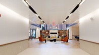Located at 27 Park Rd in West Hartford, CT, this thoughtfully designed single-floor space combines contemporary aesthetics with functional versatility. The layout flows seamlessly across several distinct zones, all unified by herringbone-patterned hardwood flooring and a cohesive modern palette.

The main lounge area serves as the heart of the space, featuring soaring double-height ceilings accented by bold navy blue architectural elements and a striking sculptural chandelier. Comfortable seating arrangements—including plush sofas, accent chairs in warm terracotta tones, and coffee tables—are anchored by a large abstract area rug, creating an inviting and social atmosphere. Built-in bookshelves flank a central fireplace, adding warmth and character.

The entry corridor features sleek recessed linear lighting and glass-paneled doors, leading naturally into the open lounge. A designated dining nook with tufted leather banquette seating and marble-top tables provides an intimate setting for gatherings.

A private meeting room offers a refined environment with warm wood paneling, a marble conference table, and a large wall-mounted display screen. The kitchen area is a standout feature, showcasing navy cabinetry, a generous marble-topped island with wood bar stools, pendant lighting, and gold-toned ceiling details, delivering both style and practicality.