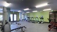 This fitness center offers a thoughtfully designed, single-floor workout space ideal for a comprehensive exercise routine. The room is finished with durable rubber flooring suited for both cardio and strength training activities. Bright fluorescent lighting runs across the dark ceiling, ensuring the entire space is well-lit throughout the day. The walls are painted in a fresh sage green, contributing to an energizing yet comfortable atmosphere. A row of treadmills and an elliptical machine, each equipped with individual display screens, lines one side of the room, providing ample cardio options. Weight benches and additional strength training equipment are positioned throughout the remaining floor space, offering versatility for a range of workout routines. A wall-mounted mirror near the rear of the room enhances the sense of openness and supports proper form during exercise. French doors along one wall allow natural light to filter in and provide easy access to the exterior. A clearly marked emergency exit ensures safety compliance. The overall layout is open and functional, making efficient use of the available space while maintaining clear pathways between equipment.