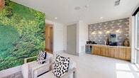 Located in Winter Garden, Florida, this thoughtfully designed communal lounge space offers a welcoming and visually dynamic environment across a single floor. The space is organized into a living area, dining area, and connecting hallway, each contributing to a cohesive and modern aesthetic.

The living room serves as the centerpiece, anchored by a striking floor-to-ceiling botanical moss wall mural that introduces a bold natural element against the otherwise crisp white walls and light tile flooring. Comfortable linen-upholstered armchairs and a wood-framed coffee table are arranged around a textured area rug, while an oversized woven pendant light adds warmth and character overhead.

Adjacent to the seating area, a compact kitchenette features warm wood cabinetry, a beverage refrigerator, and a geometric patterned accent wall in soft grey tones — ideal for a coffee or refreshment station.

The dining area offers a casually elegant atmosphere, with a channeled upholstered banquette running beneath a blue textured accent wall. White tulip-style pedestal tables paired with sleek black chairs provide ample seating, all illuminated by pendant lights with a mid-century silhouette. Floor-to-ceiling glass windows flood the dining nook with natural light and offer open views of the surrounding Florida landscape, enhancing the airy, resort-inspired ambiance throughout.