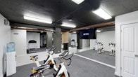 This single-floor fitness center offers a comprehensive workout environment designed to accommodate a variety of exercise preferences and training styles. The space is anchored by a row of True-brand stationary cycling bikes positioned on durable carpet flooring, ideal for high-energy spin sessions. A large wall-mounted mirror runs along one side, enhancing the sense of openness while allowing users to monitor their form during workouts. A flat-screen display mounted on the mirror wall features Fitness on Demand programming, providing guided on-demand classes directly within the space. Treadmills and additional cardio equipment are visible toward the back of the room, offering further training options. Exposed pipes and structural columns punctuate the ceiling, lending the space an industrial aesthetic that is complemented by the dark-painted ceiling and overhead fluorescent lighting. A secondary area is visible in the background, suggesting dedicated zones for strength or functional training. The overall layout is practical and efficient, making thoughtful use of the available floor space to deliver a well-rounded fitness facility.
