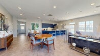 Located at 201 Idlewild Rd in Bel Air, Maryland, this single-floor community space offers a thoughtfully designed open-plan layout that seamlessly integrates a living room, dining area, and full kitchen. The kitchen is a standout feature, showcasing bold navy blue shaker-style cabinetry that extends to the ceiling, complemented by granite-topped counters and a generously sized island with bar seating. Globe pendant lights hang overhead, while stainless steel appliances — including a French door refrigerator, dishwasher, and microwave — complete the well-appointed space. The herringbone-patterned tile flooring flows throughout, lending a refined, cohesive aesthetic. The living room is bright and inviting, benefiting from abundant natural light pouring through large multi-pane windows that frame outdoor views of the surrounding landscape. A corner stacked-stone fireplace with a classic white mantle serves as a focal point, adding warmth and character to the room. The dining area offers comfortable seating around round wooden tables, ideal for gatherings, and opens to the main corridor via sliding glass doors. Soft gray walls, recessed lighting, and curated artwork throughout create a polished, welcoming ambiance suited for community use.