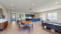 Located at 201 Idlewild Rd in Bel Air, Maryland, this single-floor community space offers a thoughtfully designed open-plan layout that seamlessly integrates a living room, dining area, and full kitchen. The kitchen is a standout feature, showcasing bold navy blue shaker-style cabinetry that extends to the ceiling, complemented by granite-topped counters and a generously sized island with bar seating. Globe pendant lights hang overhead, while stainless steel appliances — including a French door refrigerator, dishwasher, and microwave — complete the well-appointed space. The herringbone-patterned tile flooring flows throughout, lending a refined, cohesive aesthetic. The living room is bright and inviting, benefiting from abundant natural light pouring through large multi-pane windows that frame outdoor views of the surrounding landscape. A corner stacked-stone fireplace with a classic white mantle serves as a focal point, adding warmth and character to the room. The dining area offers comfortable seating around round wooden tables, ideal for gatherings, and opens to the main corridor via sliding glass doors. Soft gray walls, recessed lighting, and curated artwork throughout create a polished, welcoming ambiance suited for community use.