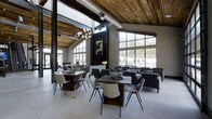 This single-story space showcases a striking blend of industrial and rustic design elements. The soaring ceiling is clad in reclaimed wood planking, complemented by exposed metal ductwork that reinforces the industrial aesthetic. A modern multi-bulb chandelier serves as a focal point overhead, adding a warm, sculptural lighting element to the voluminous interior.

Floor-to-ceiling steel-framed windows line multiple walls, allowing abundant natural light to pour in while offering views of the surrounding landscape. The large-format light-toned tile flooring contrasts beautifully with the warm wood tones above, creating a balanced visual dynamic throughout the space.

The open floor plan is furnished with a mix of rustic wood dining tables paired with contemporary woven and upholstered chairs, alongside clusters of modern lounge seating. A dark-framed fireplace anchors one wall, adding a cozy focal point amid the expansive layout. Artwork displayed above the fireplace adds a curated, thoughtful touch. The overall ambiance is sophisticated yet welcoming, merging raw industrial materials with refined furnishings.