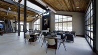 This single-story space showcases a striking blend of industrial and rustic design elements. The soaring ceiling is clad in reclaimed wood planking, complemented by exposed metal ductwork that reinforces the industrial aesthetic. A modern multi-bulb chandelier serves as a focal point overhead, adding a warm, sculptural lighting element to the voluminous interior.

Floor-to-ceiling steel-framed windows line multiple walls, allowing abundant natural light to pour in while offering views of the surrounding landscape. The large-format light-toned tile flooring contrasts beautifully with the warm wood tones above, creating a balanced visual dynamic throughout the space.

The open floor plan is furnished with a mix of rustic wood dining tables paired with contemporary woven and upholstered chairs, alongside clusters of modern lounge seating. A dark-framed fireplace anchors one wall, adding a cozy focal point amid the expansive layout. Artwork displayed above the fireplace adds a curated, thoughtful touch. The overall ambiance is sophisticated yet welcoming, merging raw industrial materials with refined furnishings.