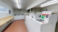 This communal laundry facility is a practical, well-organized space designed to accommodate multiple users simultaneously. The room features a row of top-loading washing machines prominently positioned along the central and side areas, complemented by a set of dryers along the back wall, providing ample capacity for laundry needs. A utility sink with exposed faucets is conveniently installed within the space, offering added functionality for hand-washing or pre-treating garments. A built-in folding table along one wall provides a dedicated surface for sorting and folding laundry. The flooring consists of durable terracotta-toned ceramic tiles, well-suited for a high-traffic utility environment. The walls are finished in a neutral sage green tone, contributing to a clean and functional aesthetic. Overhead fluorescent lighting ensures the space is brightly and evenly illuminated throughout. Low-profile dividers help organize the layout, creating a sense of order within the room. Overall, the facility is designed with efficiency and practicality in mind, offering residents a clean, functional, and well-maintained environment for their laundry needs.