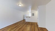 This single-floor space offers a clean, open layout defined by warm-toned hardwood flooring that runs throughout, lending a sense of continuity and warmth. One of the most distinctive architectural details is the white-painted brick accent wall, which adds texture and character to the interior while maintaining a bright, airy feel alongside the white walls and ceiling.

The main living area is open and well-proportioned, receiving natural light from a window visible toward the rear of the space. Flush-mount ceiling light fixtures provide consistent overhead lighting throughout. A compact pass-through counter connects the living area to the kitchen, allowing for an open sightline while maintaining a defined cooking zone. The kitchen features a white refrigerator and is neatly tucked into its own area.

An electrical panel is mounted on the wall near the doorway, and a visible door leads to an adjacent room. The sloped ceiling detail near the kitchen adds a subtle architectural element. Overall, the space feels straightforward, functional, and versatile, with classic materials and a neutral palette that offer a strong foundation for a variety of uses.