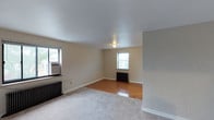 This single-floor space offers a straightforward and functional layout centered around a spacious main living area. The room features a combination of warm parquet hardwood flooring and carpeted sections, adding visual interest and texture underfoot. Neutral beige walls and white ceilings create a clean, understated backdrop throughout.

Two windows allow natural light to filter into the space, contributing to a bright and airy atmosphere. Each window is accompanied by a cast-iron radiator beneath it, providing classic, reliable heating — a notable architectural detail that adds a touch of traditional character to the otherwise simple interior. One window is also fitted with a wall-mounted air conditioning unit, ensuring year-round climate control.

A flush-mount ceiling fixture provides overhead lighting, complementing the natural light from the windows. White baseboards trim the perimeter of the room cleanly, maintaining a tidy finish. The partial wall visible in the foreground suggests a defined transition point within the layout, hinting at additional areas beyond the main room. Overall, the space presents a practical and adaptable environment with classic residential elements.