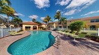 This single-story property embodies the relaxed elegance of Florida resort-style living. The standout feature of the community is its expansive freeform swimming pool, finished in vivid turquoise water and framed by a generously sized paver sundeck. The deck is well-appointed with bright green lounge chairs and umbrellas, offering ample space for sunbathing and outdoor relaxation beneath the open Florida sky.

The surrounding landscape is richly tropical, with tall swaying palm trees and lush green foliage providing a natural, resort-like ambiance. The buildings feature warm yellow stucco exteriors topped with classic terracotta tile roofing, consistent with a Mediterranean or Spanish Colonial architectural influence. An arched, open-air breezeway connects the outdoor amenity area to adjacent spaces, enhancing the indoor-outdoor flow characteristic of warm-weather communities.

White perimeter fencing borders the pool area, creating a defined and secure outdoor environment. The overall setting evokes a sense of leisurely comfort, with thoughtful landscaping and architectural details contributing to a cohesive, welcoming aesthetic. The combination of vibrant colors, tropical greenery, and well-maintained common areas makes this property a distinctive and inviting retreat suited to an active outdoor lifestyle.