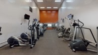 This fitness center offers a comprehensive workout environment housed within a single-level space designed to accommodate a broad range of exercise needs. The room features tall ceilings that contribute to an open, airy atmosphere, while recessed fluorescent lighting provides consistent illumination throughout. Neutral white walls are punctuated by a bold orange accent wall at one end, complemented by a large mirrored section beneath it that visually expands the space and allows users to monitor their form during exercise.

The flooring consists of durable, gray carpet throughout, suited for the demands of a high-use fitness environment. Equipment is arranged in orderly rows along both sides of the room, including elliptical trainers, treadmills, stationary bikes, and stair climbers from brands such as True, Octane, and Life Fitness. A cable strength machine and recumbent bike round out the strength and low-impact options available.

Multiple wall-mounted flat-screen televisions are positioned at eye level throughout the space, allowing users to stay entertained during cardio sessions. A small window on one wall provides a connection to the outdoors and a modest source of natural light. The overall layout is functional and well-organized, supporting an efficient and comfortable fitness experience.