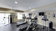 This fitness center offers a comprehensive workout environment designed to support a variety of exercise routines. The space is outfitted with multiple treadmills and an elliptical machine, providing ample cardio options for users. A full dumbbell rack stocked with a wide range of free weights sits along one wall, catering to strength training needs. A cable machine and additional resistance equipment round out the functional training options available.

The interior features neutral beige walls with white trim and recessed lighting complemented by a ceiling fan, ensuring the space remains well-lit and comfortable during use. Multiple wall-mounted flat-screen televisions are positioned throughout the room, allowing members to stay entertained while exercising. A window along one wall allows natural light to filter in, adding to the open and energizing atmosphere of the space.

The dark-toned flooring provides a practical and durable surface suited for fitness use. A wall-mounted clock keeps exercisers on track with their workout timing. The overall layout is thoughtfully arranged to maximize usability while maintaining clear pathways between equipment, creating a functional and welcoming environment for fitness enthusiasts of all levels.