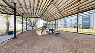This community playground is a thoughtfully designed outdoor recreational space nestled within a well-maintained residential setting. The centerpiece of the area is a generously sized open-air pavilion with a metal truss roof supported by steel columns, providing ample shade and weather protection for year-round play. Beneath the covered structure, a multi-component play system features slides, climbing walls, and various activity panels, all set atop a cushioned wood chip surface bordered by durable edging for safety and containment. The open-sided design of the pavilion allows for natural airflow and clear sightlines, making supervision easy and the environment feel open and inviting. Surrounding the pavilion, the landscape is lush and well-kept, with manicured green lawns, mature palm trees, and ornamental plantings lining curved mulched garden beds. Smooth concrete walkways wind through the grounds, connecting the playground to adjacent outdoor seating areas and covered patios attached to the neighboring residential buildings. The overall ambiance is clean, family-friendly, and community-oriented, with the residential buildings visible in the background, reinforcing the sense of a cohesive and active neighborhood environment.