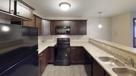 This single-floor unit is designed with a functional and cohesive aesthetic throughout its living spaces. The kitchen serves as a central highlight, showcasing rich dark wood cabinetry that contrasts elegantly with light granite-look countertops. Black appliances, including a refrigerator, electric range, and over-the-range microwave, lend a sleek, uniform appearance. The U-shaped kitchen layout maximizes counter and storage space, while wood-look vinyl flooring adds durability and a modern touch. Pendant and flush-mount lighting illuminate the space effectively.

The bedrooms feature neutral-toned carpet flooring and large windows fitted with blinds, allowing for natural light while maintaining privacy. Each room offers a clean, versatile canvas with ample wall space and floor vents for climate control.

The bathroom is finished in a neutral gray palette, featuring a single-sink vanity with a wide mirror, a tub-shower combination, and notably, an in-unit washer and dryer tucked conveniently into the same space. Overall, the unit presents a practical and well-maintained interior with consistent finishes throughout.