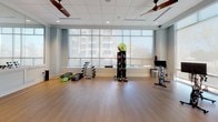 Located at 1108 S Main St in Greenville, South Carolina, this single-floor fitness center offers a well-appointed and thoughtfully designed workout environment. The space is characterized by soft blue-gray walls, warm hardwood-style flooring, and a drop ceiling with recessed lighting and ceiling fans that promote air circulation throughout.

The main workout area is open and spacious, providing ample room for a variety of exercise activities. A floor-to-ceiling mirror wall runs along one side, visually expanding the space and allowing users to monitor their form during workouts. Large, floor-to-ceiling windows line the perimeter, flooding the interior with abundant natural light and offering views of the surrounding Greenville streetscape.

A dedicated cycling zone features a row of stationary bikes equipped with individual screens, positioned directly in front of the expansive windows for an energizing workout experience. Adjacent to this area, a well-stocked free weight section includes a dumbbell rack, step platforms, kettlebells, and medicine balls organized on a standalone tower.

One wall is equipped with a large mounted flat-screen display alongside a smaller tablet interface, offering on-demand fitness content and workout guidance. A mirrored partition elegantly separates zones within the facility while maintaining an open, connected feel. The overall ambiance is clean, modern, and inviting, designed to support a full range of fitness activities.