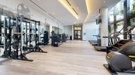 Located at 11295 Biscayne Blvd in Miami, FL, this contemporary fitness center offers a well-appointed, single-floor layout designed to support a full range of workout routines. The main gym area is expansive and generously lit, with floor-to-ceiling windows lining one side that frame lush tropical palm views and invite abundant natural light throughout the space. Light hardwood-style flooring runs throughout, complementing the clean, modern aesthetic defined by crisp white walls, recessed lighting, and a curved tray ceiling with warm ambient uplighting that adds a refined architectural touch. The main floor is equipped with a comprehensive selection of commercial-grade cardio machines — including treadmills, ellipticals, stair climbers, and rowing machines — all positioned to take advantage of the outdoor views. A dedicated strength training zone features cable machines, power racks, weight benches, and free weights, accented by a decorative perforated wall panel that adds visual interest. A separate enclosed room serves as a specialized boxing and functional training studio, outfitted with an Everlast punching bag system, medicine balls, a stability ball, and a training mat, all under soft pendant lighting. The overall ambiance is bright, clean, and motivating — a premium wellness environment befitting its Miami setting.