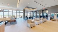Located at 9600 S Dixie Hwy in Miami, FL, this single-floor amenity space is a thoughtfully designed, multi-functional environment that blends work, leisure, and outdoor living seamlessly. Floor-to-ceiling glass walls and expansive window systems flood the interior with abundant natural light while offering sweeping views of the surrounding Miami skyline and lush greenery.

The open-plan interior is divided into distinct yet connected zones. A dedicated coworking area features a central conference table with ergonomic seating, pendant lighting, and private glass-enclosed offices along the perimeter — ideal for focused work. Adjacent to this, a social lounge area offers modular white upholstered seating arranged for casual gathering and collaboration.

A warm wood-accented media wall with an integrated flat-screen TV and dark marble countertop anchors a comfortable lounge and dining zone, complemented by large-format porcelain tile flooring throughout. Concrete cylindrical columns add an industrial architectural accent.

The entertainment zone includes classic arcade cabinets — PAC-MAN and PONG — alongside blue velvet seating, bringing a playful energy to the space. Large sliding glass doors open directly onto a lushly landscaped outdoor terrace featuring artificial turf, hanging swing chairs, lounge seating, a punching bag, and pergola-covered gathering areas — extending the living space into the outdoors.