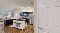 Located at 13 Hawthorne Gate Dr in Dayton, Ohio, this single-floor home offers a well-designed, open-concept layout with rich hardwood-style flooring that flows throughout the main living areas. The kitchen is a standout feature, showcasing dark espresso cabinetry with granite countertops, a full suite of stainless steel appliances, and a contrasting white island with a dark countertop and built-in storage. A dedicated walk-in pantry provides generous shelving for organized storage. Pendant lighting and recessed fixtures illuminate the space, which opens seamlessly into the living room. The living area features vaulted ceilings and a warm, inviting ambiance with natural light filtering through large windows and a sliding glass door. The bedroom is carpeted for comfort and is served by a ceiling fan and a window with sheer drapery. A dedicated home office features hardwood-style flooring, a ceiling fan, and a bright window, offering a functional workspace. Two full bathrooms are finished with dark cabinetry, granite-topped vanities, and hardwood-style flooring. One bathroom includes an accessible grab bar and in-unit washer and dryer, adding everyday convenience. The overall design is cohesive, modern, and practical throughout.