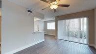 This single-story space showcases a bright, open-concept layout designed for comfortable, everyday living. The main living area features smooth wood-style laminate flooring in a neutral gray tone, complementing the soft beige walls and crisp white trim throughout. A ceiling fan with an integrated light fixture is centrally mounted on the textured popcorn ceiling, providing both airflow and ambient lighting.

A defining architectural feature is the pass-through kitchen counter, which opens directly into the living area. This half-wall design creates a seamless visual connection between the kitchen and living space, with a modern pull-down faucet and sink visible at the counter level. Track lighting above the pass-through adds focused illumination to the workspace. A window adjacent to the counter allows additional natural light to filter into the kitchen area.

The living space is anchored by a large sliding glass door dressed with vertical blinds, which floods the interior with natural light and provides access to an outdoor area. The overall ambiance is clean, modern, and move-in ready, with a neutral palette that offers versatility for a variety of furnishing styles.