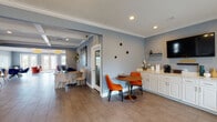 This well-appointed single-story space is designed with a cohesive mid-century modern aesthetic, characterized by cool blue-gray walls, crown molding, and wood-look tile flooring that flows throughout. The entry foyer sets an inviting tone with a statement chandelier, a large mirror flanked by geometric wall art, and a pair of plush white accent chairs, creating a polished first impression.

Moving further inside, a reception area features a rustic wood desk set against a striking floor-to-ceiling mosaic tile accent wall, adding texture and visual depth. Adjacent to this is a compact refreshment station with white cabinetry, a marble-style countertop, and a coffee setup, alongside a vibrant round dining table paired with bold orange chairs.

A separate meeting or lounge area offers a round wood table with cobalt blue chairs, open shelving with greenery, and direct access through French doors to an adjoining space. The rear lounge is a standout zone, with terracotta velvet chairs, a colorful geometric area rug, and a wall of French doors that flood the room with natural light and frame expansive views of the pool deck and palm trees beyond, creating a bright, resort-like ambiance.