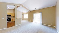 This single-floor home offers a thoughtfully designed open-concept layout that maximizes both space and natural light. The living area is anchored by striking vaulted ceilings with exposed beam detailing, creating an airy, expansive atmosphere throughout. Warm neutral tones on the walls complement the plush wall-to-wall carpeting underfoot, lending the space a welcoming and comfortable feel.

A defining feature of the main living area is the sliding glass door that opens to a private outdoor deck, framing views of mature trees and allowing natural light to pour into the room. The seamless connection between indoor and outdoor spaces enhances the overall sense of openness.

The kitchen is partially separated from the living area by a half-wall pass-through with a countertop bar, offering both functional workspace and an informal dining option. Natural wood cabinetry is visible in the kitchen, adding warmth and classic character. A pendant light fixture hangs above the dining area just beyond the pass-through, defining the space with a touch of elegance.

The interplay of vaulted ceilings, multiple windows, and the glass door ensures the home is bathed in natural light throughout the day, making this a bright and inviting single-floor residence suited for comfortable everyday living.