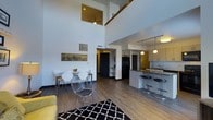 This two-story space makes an immediate impression with its dramatic double-height ceilings in the main living area, creating a sense of volume and openness that defines the entire lower floor. The open-concept layout seamlessly connects the living, dining, and kitchen zones, encouraging a natural flow between spaces. Hardwood-style flooring runs throughout, anchoring the modern aesthetic.

The kitchen is well-appointed with light wood cabinetry, a dark countertop peninsula, and a combination of pendant and track lighting that provides both task and ambient illumination. Bar-height stools line the peninsula, making it a functional spot for casual dining or conversation.

The dining area features a compact table with modern white chairs, positioned beneath the soaring ceiling and adjacent to the living space. A black-and-white patterned area rug defines the living zone, complemented by a yellow accent chair that adds a pop of color.

A second floor is visible from the main level, with a railing overlooking the open living area below and a ceiling fan adding to the airy atmosphere. The overall ambiance is contemporary and bright, with a clean, urban feel throughout.