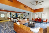 Located in Tewksbury, MA, this lobby and lounge offers a generously proportioned, single-floor communal space designed with both comfort and style in mind. The interior is immediately distinguished by its bold orange ceiling accents set against neutral gray-white walls, creating a vibrant yet welcoming atmosphere. A prominent stone fireplace serves as a focal point in the lounge area, framed by large arched windows that flood the space with natural light and provide views of the outdoors. Ceiling fans overhead contribute to a comfortable environment throughout.

The main lounge features tufted cognac leather sofas arranged around a round coffee table atop a blue geometric-patterned carpet, ideal for casual gatherings. A secondary seating cluster with striped armchairs is positioned near the front reception desk, which is finished in warm wood tones and anchors the entry vestibule. The dining area includes a dark oval table surrounded by plush orange upholstered chairs, illuminated by sculptural globe pendant lights, with framed artwork adorning the walls and floor-to-ceiling windows offering outdoor views. Blue accent columns define the transition between zones, while curated wall art and greenery add finishing touches throughout the space.