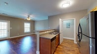 This single-floor unit offers a thoughtfully designed open-concept layout that seamlessly connects the kitchen and living areas. The kitchen serves as a functional centerpiece, featuring a peninsula with a double sink and neutral countertops, warm-toned wood cabinetry extending to the ceiling, and a full suite of stainless steel appliances including a refrigerator, electric range, built-in microwave, and dishwasher. Rich hardwood-style flooring flows throughout the main living areas, adding warmth and continuity to the space.

The living area is generously proportioned, with crown molding accents, a ceiling fan with light fixture, and large windows that allow natural light to fill the room. A glass panel door provides access to the outdoors, further brightening the space. The bedroom continues the comfortable aesthetic with plush carpeting, a ceiling fan, and a window offering views of the surrounding greenery.

The bathroom is clean and functional, equipped with a full-size soaking tub and shower combination featuring chrome fixtures. Additional practical elements include a utility panel accessible via a dedicated door. Neutral gray wall tones throughout the unit create a cohesive, move-in-ready atmosphere suited to a variety of furnishing styles.