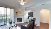 This thoughtfully designed single-floor residence offers a comfortable and stylish living experience across one bedroom and one bathroom. The living area serves as the heart of the home, highlighted by a classic white-mantled fireplace that anchors the room with warmth and character. Rich hardwood-style flooring runs throughout, complementing the crisp white walls and elegant crown molding that lend a refined, polished feel to the space. A ceiling fan with integrated lighting provides both comfort and ambiance, while decorative columns subtly define areas within the open layout. Natural light filters in through sliding glass doors fitted with vertical blinds, hinting at outdoor access. The living space is generously proportioned for its footprint, offering room for a full seating arrangement and additional accent furnishings. Architectural detailing such as the columned entryways and recessed wall niche add dimension and visual interest. The overall ambiance is bright, clean, and inviting — a balance of classic design elements and functional living that makes this residence a comfortable and attractive place to call home.