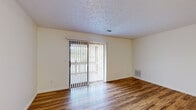This single-story space offers a well-organized layout with a variety of functional rooms finished with consistent, practical materials throughout. The main living area is a generously sized, open room featuring warm wood-look flooring that reflects natural light streaming in through a large sliding glass door fitted with vertical blinds. The sliding door provides direct access to an outdoor balcony, adding a sense of openness to the space. A textured ceiling runs throughout the common areas, contributing to the overall character of the interior.

The galley-style kitchen is efficiently arranged with rich, warm-toned wood cabinetry lining both walls, light countertops, and wood-look flooring underfoot. White appliances, including a range, microwave, refrigerator, and dishwasher, are all included. A dedicated utility room houses the water heater, electrical panel, a utility sink, and wire shelving, offering practical storage and laundry hookup space.

The bedrooms are carpeted for comfort, each featuring closets with wire shelving systems. The primary bedroom connects to an en-suite bathroom with a wood vanity, while an additional full bathroom includes a tub-shower combination, vanity with ample storage, and wood-look flooring.