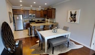 This single-floor space offers a thoughtfully designed living area that balances comfort and style. The living room serves as the heart of the space, showcasing rich hardwood flooring that adds warmth and character throughout. Crown molding accents the walls and ceilings, lending a refined, polished look to the interior.

A ceiling fan with integrated lighting is centrally positioned, providing both functionality and a classic aesthetic touch. The room is bathed in natural light, courtesy of a pair of elegant French doors that lead to an outdoor balcony, where views of the surrounding landscape can be enjoyed. The doors are framed with detailed white trim, adding an architectural focal point to the space.

The living area is generously sized, comfortably accommodating a plush sofa, a round wood coffee table, and a patterned area rug. The neutral gray wall palette creates a versatile backdrop, complemented by bold decorative artwork. The overall ambiance is relaxed yet sophisticated, making it well-suited for both everyday living and entertaining. The seamless connection between the interior and the outdoor balcony enhances the sense of openness and brings a refreshing flow of natural light into the space.